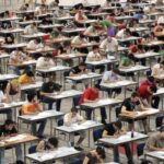 Aerial view of a large exam hall with hundreds of students at individual desks taking tests.