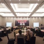 Stage with panel of speakers and a maroon banner at the front; attendees seated at round banquet tables in a well-lit conference hall.