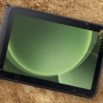 Rugged tablet lying on dirt ground, screen showing green abstract circular graphics.
