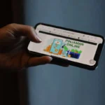 Hand holding a smartphone displaying a graphic with 'PINJAMAN ONLINE' and money icons, suggesting online loans.