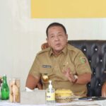 Man in a tan uniform seated at a desk, gesturing with his right hand as he speaks.