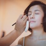 Makeup artist applies eye makeup to a woman with eyes closed and short dark hair.
