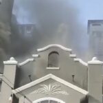 Gray ornate building in the foreground with thick black smoke rising from behind it, indicating a fire in the background.
