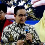 Man in patterned shirt speaks into a handheld microphone at a public event, flanked by a man in a red blazer and a woman in a pale yellow hijab.