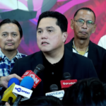 Spokesperson in a black suit speaks at a press conference, reporters surround him with multiple microphones and phones nearby.