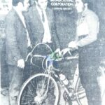 Three men stand around a vintage bicycle with a BBC sign in the background.