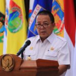 Official in white uniform giving a speech at a podium with a microphone, flags in the background.