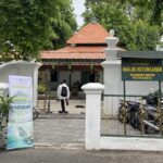 Entrance of Masjid Rotowijayan Kraton Yogyakarta: white walls, red-tiled roof, decorative gate, and a sign near parked motorcycles under leafy trees.