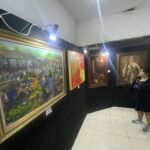 Woman in a black dress and sneakers taking a photo inside a small art gallery with black walls and white tiled floor, several framed paintings on display.