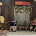 Two people sit on ornate carved chairs on a decorated stage; a large drum stands to the left and a flower garland arches over a draped backdrop with a red sign above.