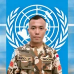 Soldier in camouflage with Indonesia flag patch standing against a United Nations emblem backdrop.