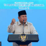 Indonesian official in beige uniform and black cap speaks at a podium with national emblem, gesturing with a raised fist during a groundbreaking ceremony backdrop reading '13 Proyek Hilirisasi Nasional Tahap II'