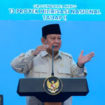 Indonesian official in beige uniform and peci speaks at a podium with the national emblem; blue backdrop reads '13 Proyek Hilirisasi Nasional Tahap II'.