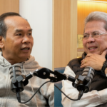 Two men in a podcast studio sit across from each other, speaking into shared microphones.
