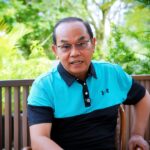 Man wearing a turquoise and black polo shirt sits on a wooden chair outdoors with lush green plants in the background, speaking or posing for a photo.