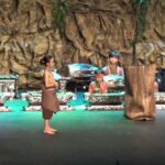 Group of young performers in brown costumes standing on a stage with a rocky backdrop, listening to a speaker on the right.