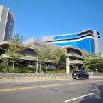 Modern multi-story building with blue glass facade along a busy street, with cars and motorcycles.
