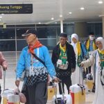 Group of travelers with rolling suitcases walking through an airport terminal, signposted for boarding lounge above them.