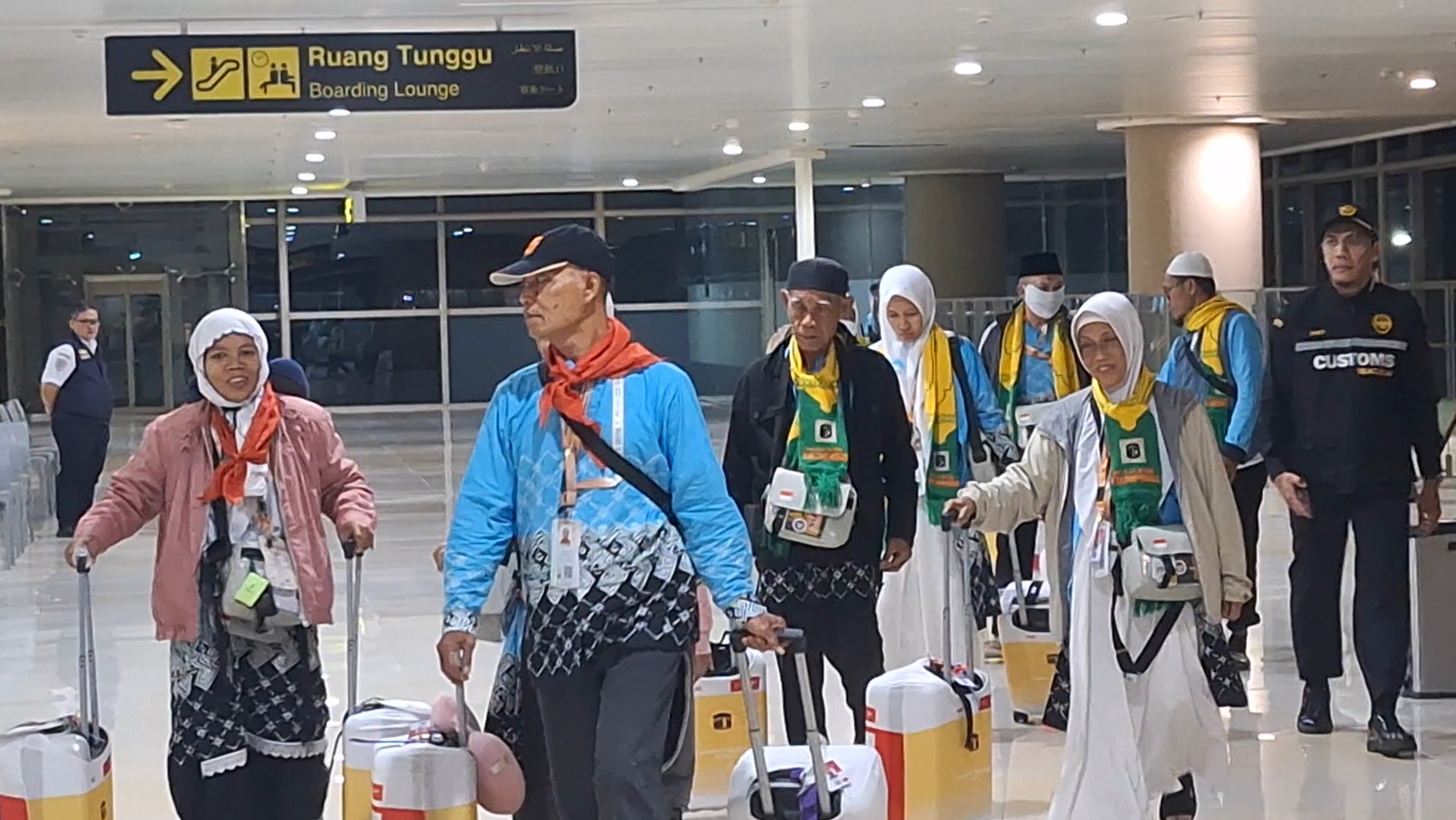 Group of travelers with rolling suitcases walking through an airport terminal, signposted for boarding lounge above them.