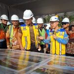Group of construction workers and officials in hard hats and safety vests gathered around a large table map while a man with a microphone explains.
