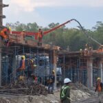 Construction workers in yellow hard hats and orange vests stand on scaffolding as a concrete pump pours into a building frame.