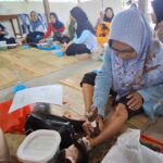 Group of women in hijabs seated on a mat in a circle, participating in a foot care workshop; a woman at the front applies product to a participant's foot.