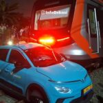 Blue SUV with a 'GREEN SM' logo sits on tracks next to a stationary orange-and-gray train at night, red emergency lights flashing and onlookers nearby.