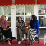 Three women in hijabs seated on a small stage outdoors; one speaks into a microphone while the others listen, under a red canopy.