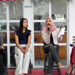 Panel of four women seated indoors; the second woman from left listens as the hijab-wearing speaker raises a microphone.