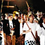 Night-time procession with young men in ceremonial attire holding staffs and banners.