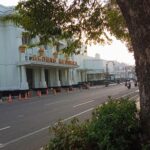 Illustration of a white building labeled 'Gedung Merdeka' beside a tree-lined street with orange traffic cones along the curb.