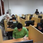 Computer lab with individual cubicles; people work at desktop computers, some wearing hijabs. A woman leans over a workstation in the foreground.