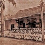 Small group photo of students in uniforms posing in front of a long traditional building with palm trees nearby.