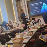 Group of professionals in hijabs seated around a table, listening to a presenter in a conference room with a blue pyramid slide on screen behind them.