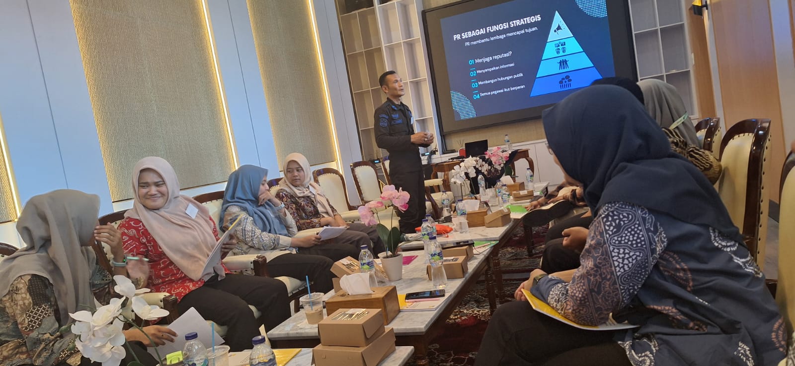 Group of professionals in hijabs seated around a table, listening to a presenter in a conference room with a blue pyramid slide on screen behind them.