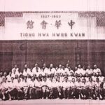 Group photo of dozens of people in white shirts posing in front of a building with a sign displaying '1907-1953' and Chinese characters; appears to be a historical organization gathering.