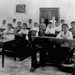 Black-and-white photo of young students seated at wooden desks in a classroom, with a teacher standing at the back near a cabinet.