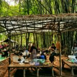 Group of people cooking and eating under a bamboo shelter with tables, baskets, and food containers in a forest setting