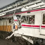 Damaged passenger train with crushed front end and shattered windows after a collision under a station canopy.