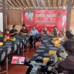 People sit around a U-shaped table in a hall; a red banner reading 'Ngobrol Bareng' dominates the background as a camera records the scene.