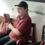 People sitting on a bench indoors, smiling and chatting; a man in a maroon polo and cap center, two women in hijabs on the left, and a man in a patterned shirt on the right. A water bottle sits on the table in front.