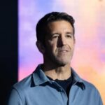 Male speaker in a blue collared shirt on stage with a colorful abstract background, delivering a talk