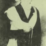 Historical black-and-white portrait of a person wearing a turban and dark robe with a light shawl, holding a bead necklace.