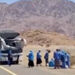 Damaged bus on a desert highway with a group of people in blue clothing nearby; rocky mountains in the background.