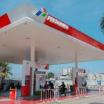 Pertamina gas station with a red-and-white canopy, fueling pumps, and motorcyclists charging at the pumps under a blue sky.