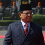 Man in a dark suit and red tie walks on a red carpet at a formal ceremony, wearing a black cap with guards in the background.