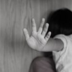 Child with an outstretched hand pressed against a wall, facing away in black and white.