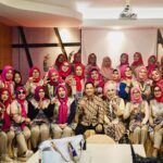 Group of women wearing pink hijabs and colorful outfits posing for a group photo at an indoor event. A few men sit in front center.