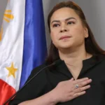 Woman speaking at a podium with her hand over her chest, the Philippine flag in the background with soft lighting.