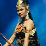 Female performer in an ornate gold headpiece and red/gold armor, holding a bow on a blue-lit stage background.